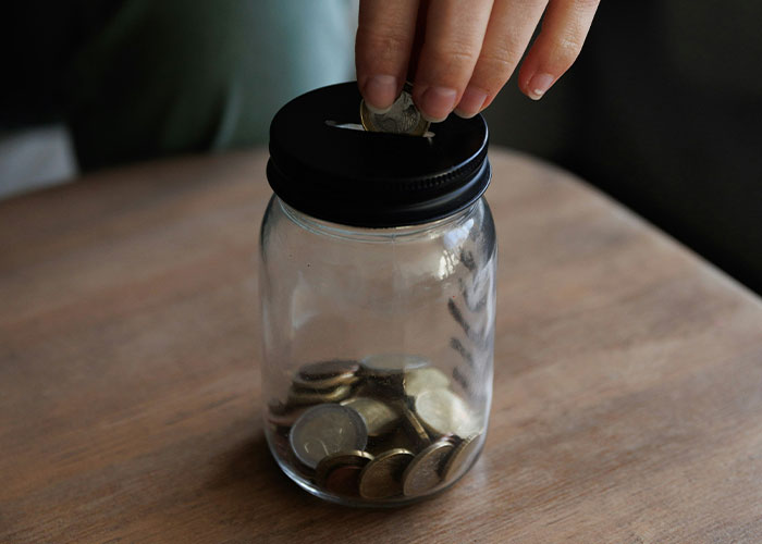 Hand putting coins into a clear jar on wooden surface, illustrating parents sharing worst kids' public moments stories.