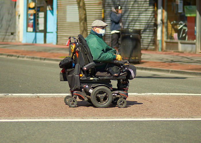 Man wearing mask and green jacket in a motorized wheelchair crossing street in a public urban area