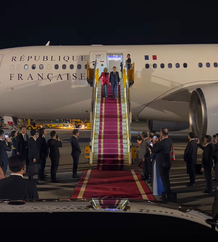 French President Emmanuel Macron and wife arriving on airplane stairs during official event with crowds watching at night.