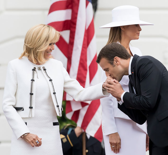 French President Emmanuel Macron kissing a woman's hand during a formal event with American flag in the background