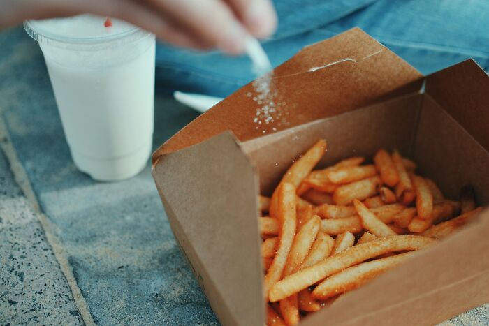 Hand sprinkling salt on French fries in a takeaway box next to a cold drink, highlighting mundane things that make people sick.