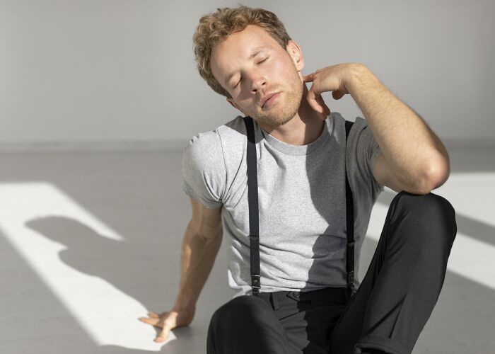 Young man with suspenders sitting on floor, looking frustrated and tired, reflecting bad habits and relationship struggles.