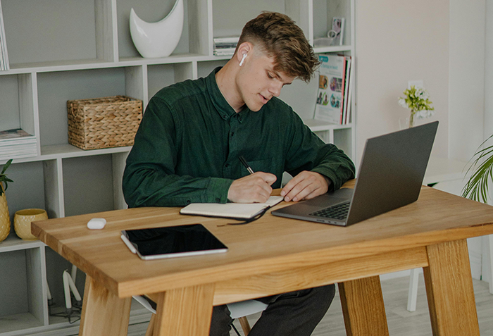 Young man working at wooden desk with laptop and tablet, writing notes, illustrating Karen fakes wellness checks concept. Young man working at wooden desk with laptop and tablet, writing notes, illustrating Karen fakes wellness checks concept.