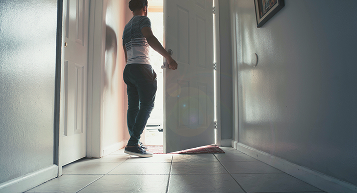 Person standing inside a house opening the front door, illustrating a wellness check or neighbor interaction scenario. Person standing inside a house opening the front door, illustrating a wellness check or neighbor interaction scenario.