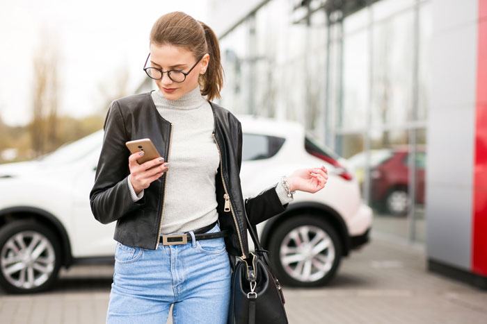 Young woman standing in a parking lot holding a phone, wearing glasses and a leather jacket near parked cars.