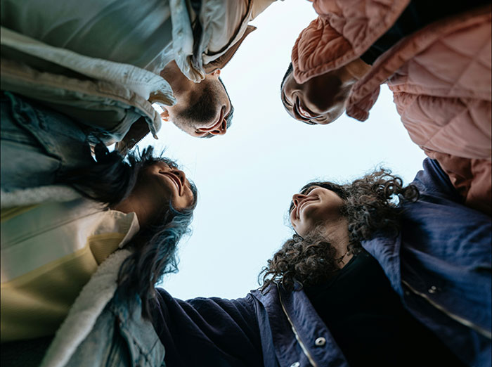 Four friends in winter jackets standing in a circle outdoors, smiling and looking down towards the camera.