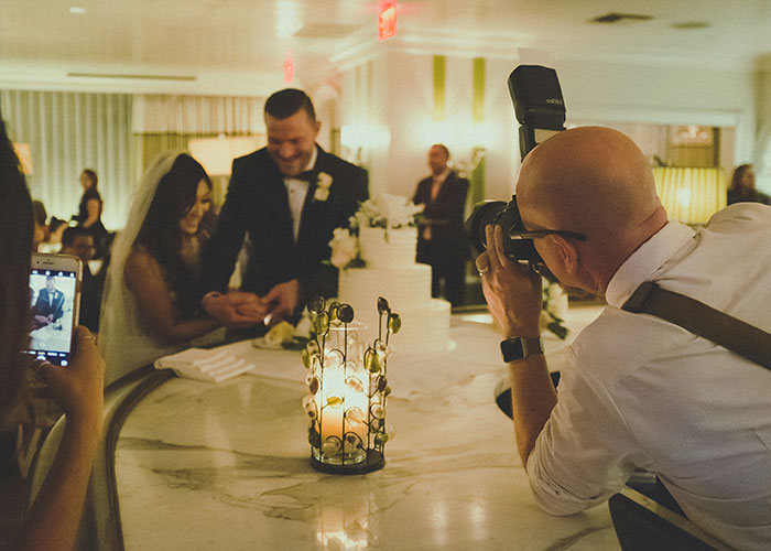 Photographer capturing a bride and groom cutting their wedding cake amid a lively celebration and guests taking photos.