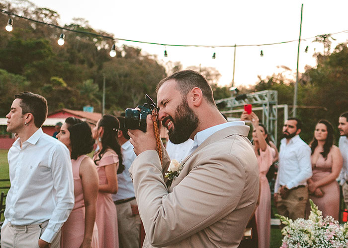 Man in beige suit taking photos at an outdoor wedding where a woman's wedding revenge sparks controversy.