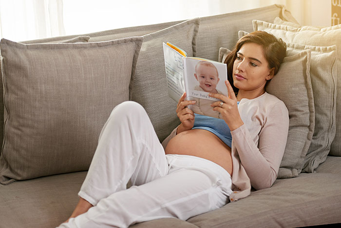 Pregnant woman lying on a couch reading a book about pregnancy and birth facts in a cozy home setting.