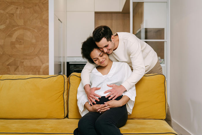 Couple embracing on yellow couch as pregnant woman sits smiling, highlighting wedding venue booking and family surprises.