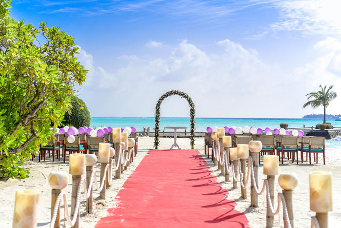 Beach wedding venue decorated with chairs, an arch, and a red carpet aisle under a clear blue sky.