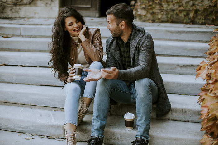 A man and woman sitting on outdoor steps, sharing coffee and smiling while having a friendly conversation.