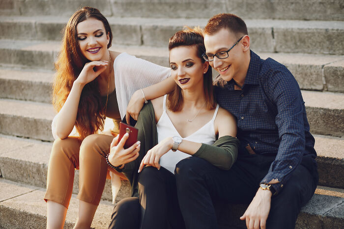 Three friends sitting on steps, with men and women capturing moments on a smartphone, highlighting lost friendships.