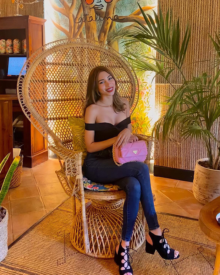 Young woman seated in a wicker chair at a vacation hotspot, smiling and holding a pink purse amid tropical decor. Young woman seated in a wicker chair at a vacation hotspot, smiling and holding a pink purse amid tropical decor.