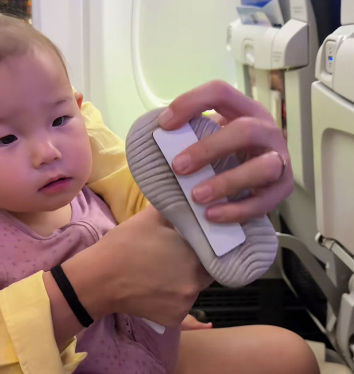 Baby&rsquo;s shoe with adhesive being attached to airplane seat back during flight as a parent holds the child nearby.