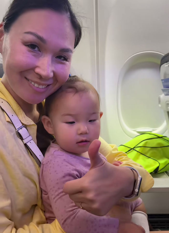 Mom holding baby on airplane, showing thumbs up, highlighting baby shoes stuck to seat back during flight controversy.