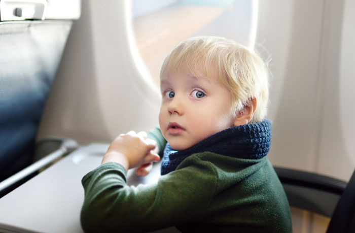 Toddler sitting in airplane seat by window, wearing a green sweater and navy scarf, looking back with curious expression.