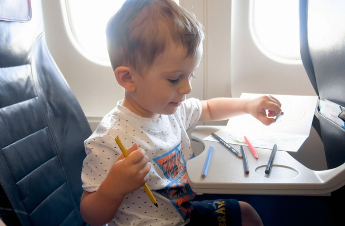 Toddler sitting in airplane seat, coloring on tray table with colored pencils during flight, illustrating baby travel tips.
