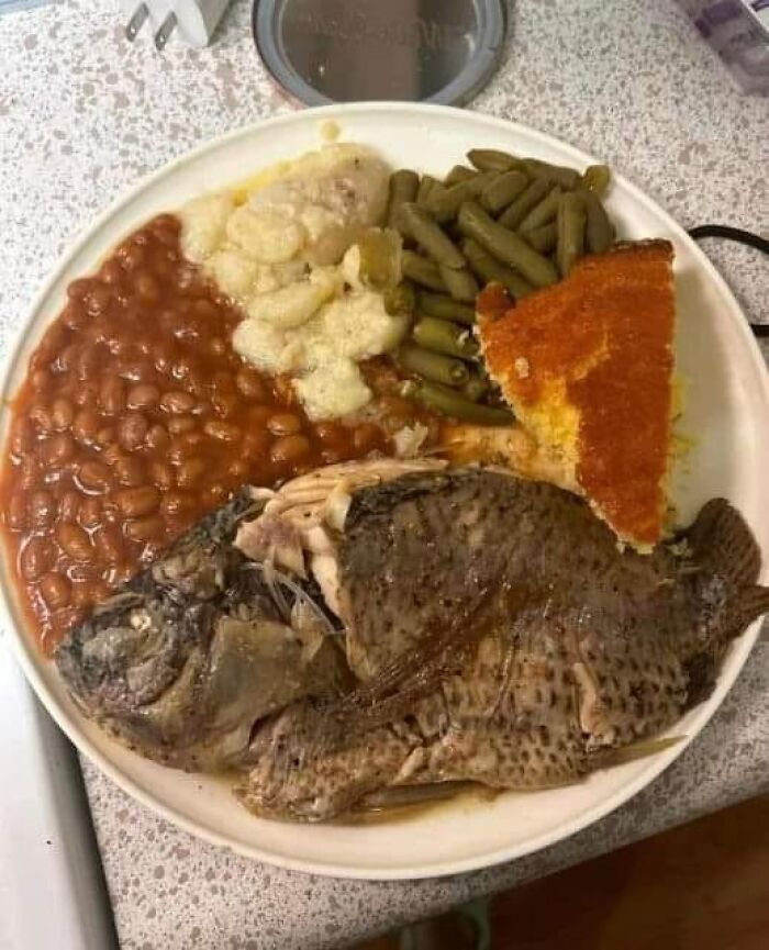 Plate of baked beans, overcooked fish, green beans, mashed potatoes, and cornbread in a cursed food pics style.