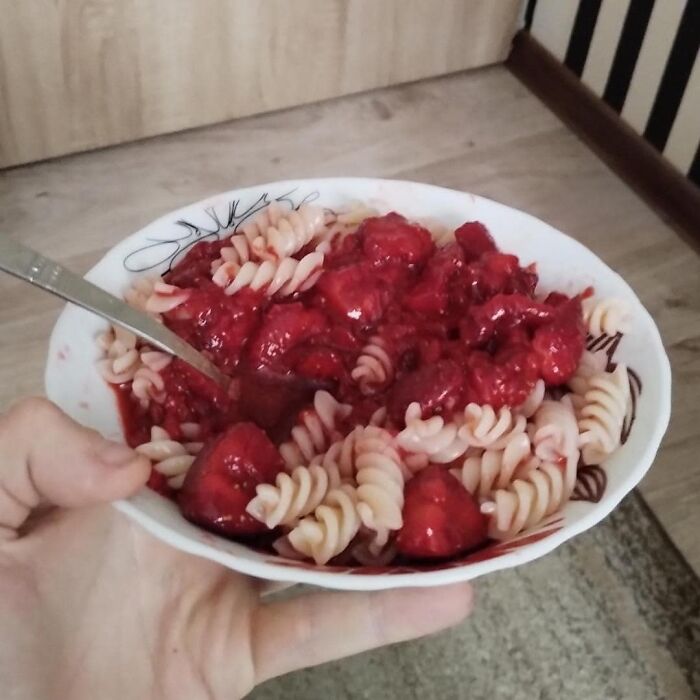 Hand holding a bowl of pasta mixed with a red sauce that looks like a cursed food pic with unusual texture.