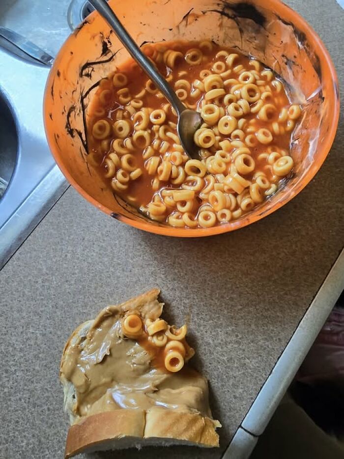 Bowl of spaghetti hoops with tomato sauce and a slice of bread spread with peanut butter topped with spaghetti in a kitchen.
