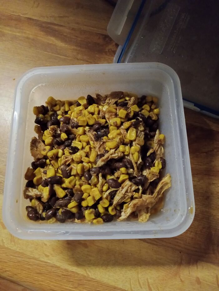 Cursed food pic of mixed black beans, corn, and shredded chicken in a plastic container on a wooden surface.