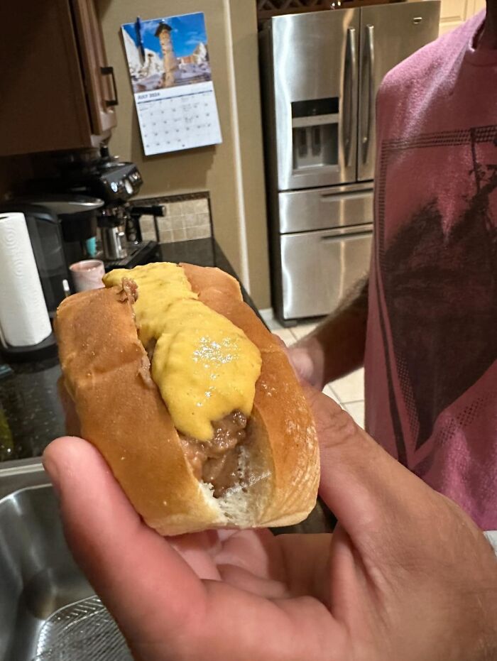 Cursed food showing a poorly made hot dog with melted cheese in a soft bun held over a kitchen sink.