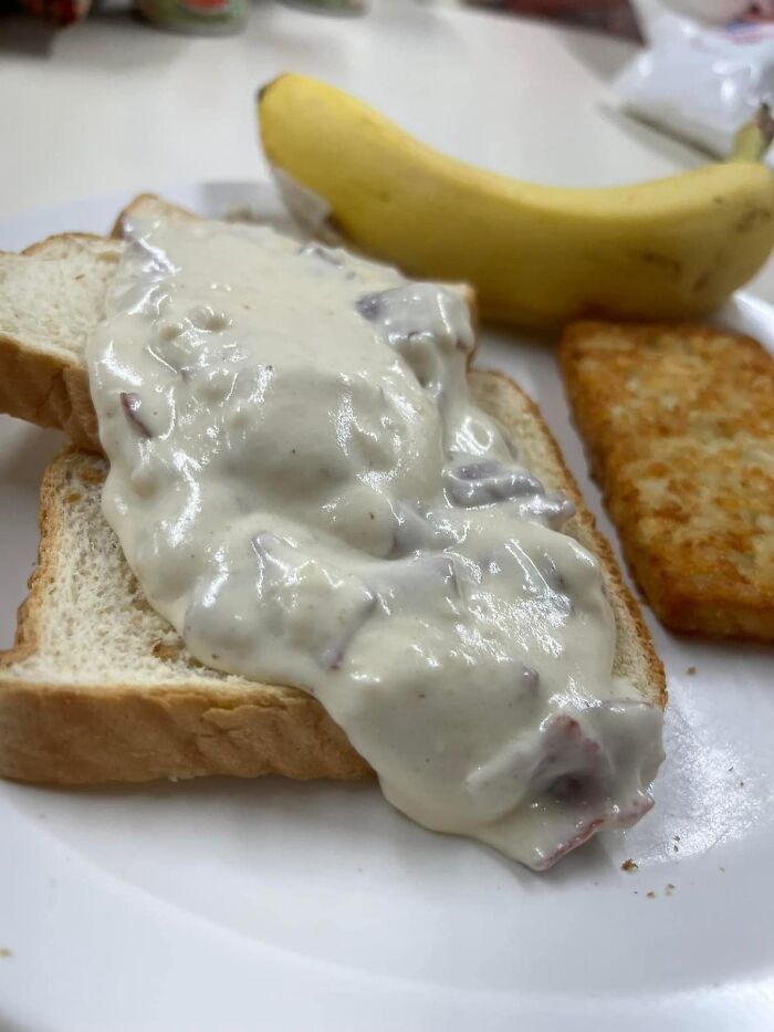 Open-faced sandwich with white gravy on bread, a whole banana, and a hash brown on a white plate, cursed food pic.
