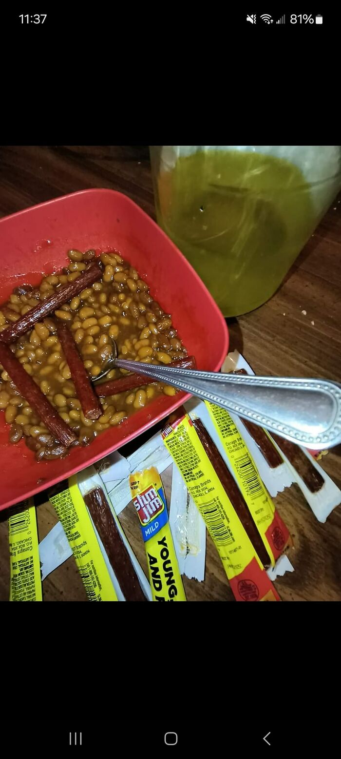 Baked beans served with hot dog sticks in a red bowl, surrounded by multiple opened Slim Jim snack wrappers on a wooden surface.