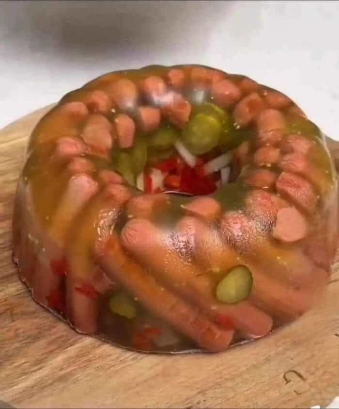 Unappetizing cursed food with hot dogs and pickles suspended in a gelatin mold on a wooden surface.