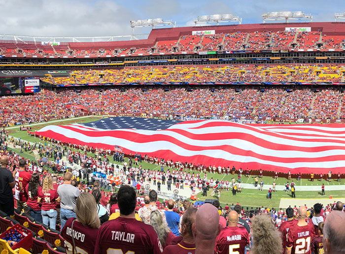 Large American flag displayed on a crowded football stadium field with fans watching during a patriotic event.