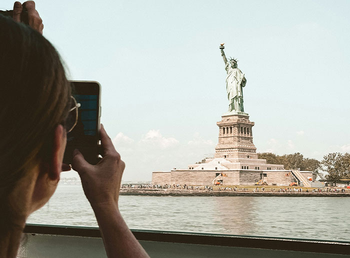 Person taking photo of the Statue of Liberty with smartphone, illustrating Americans recalling funny things foreigners said about the United States.