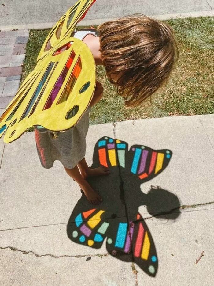 Child wearing colorful butterfly wings casting vivid butterfly shadow on sidewalk in a weird pics or it didn’t happen moment.