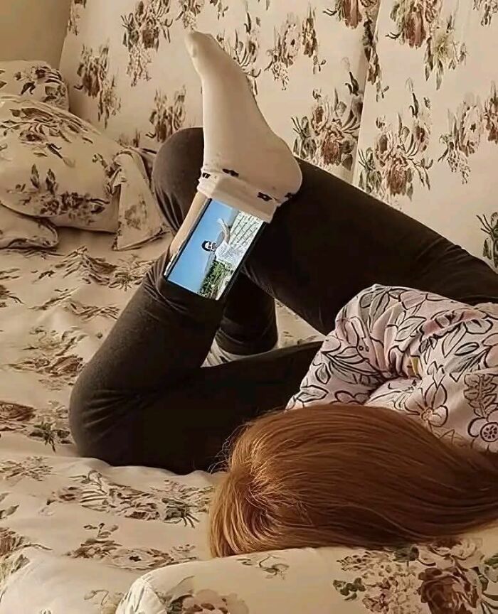 Person lying on a floral bed using a phone wedged between their socked foot and leg in a weird pics or it didn’t happen moment.