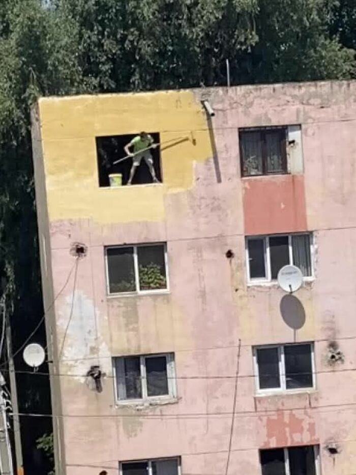 Person painting yellow wall on upper floor of worn apartment building in weird pics or it didn’t happen moments.