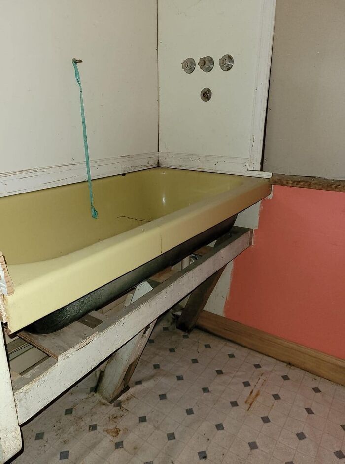 Old yellow bathtub poorly supported on wooden planks in a dirty rundown bathroom, worst real estate photos example.