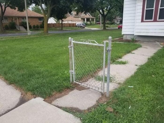 Small chain link gate detached from fence opening, awkwardly positioned on sidewalk near residential home and grass lawn.