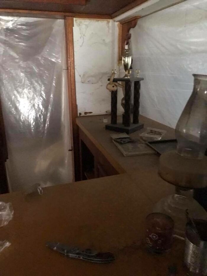 Dimly lit, cluttered room with plastic-covered walls, a trophy on a dusty counter, and various miscellaneous items scattered around.