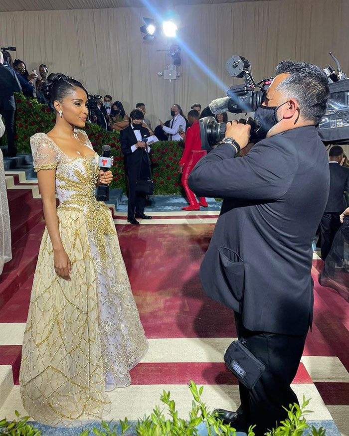 Reporter in gold embellished gown interviewed by cameraman on Met Gala red carpet with fans and celebs in background Reporter in gold embellished gown interviewed by cameraman on Met Gala red carpet with fans and celebs in background