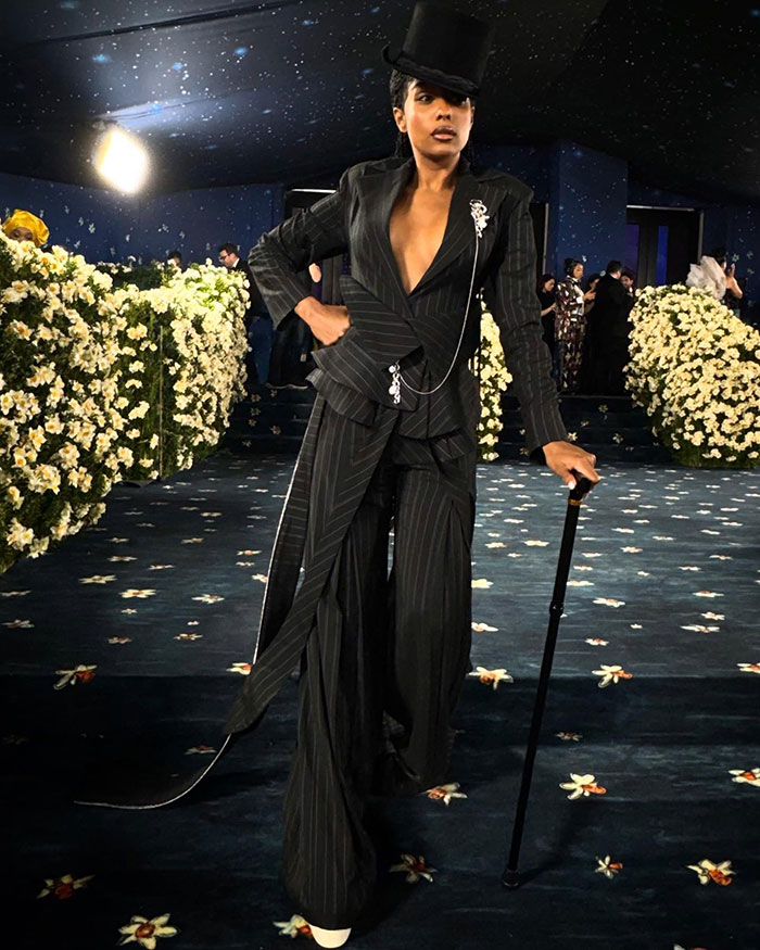 Reporter in striking pinstripe suit and top hat posing confidently with cane on Met Gala red carpet surrounded by floral decor. Reporter in striking pinstripe suit and top hat posing confidently with cane on Met Gala red carpet surrounded by floral decor.