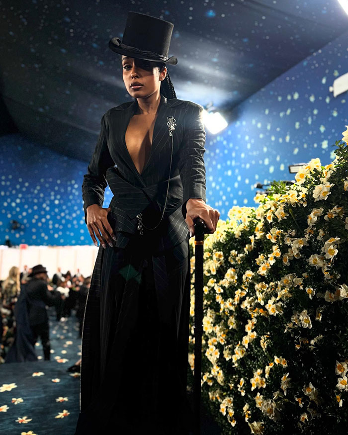 Reporter on Met Gala red carpet, wearing black pinstripe outfit and top hat, standing confidently beside floral decoration. Reporter on Met Gala red carpet, wearing black pinstripe outfit and top hat, standing confidently beside floral decoration.