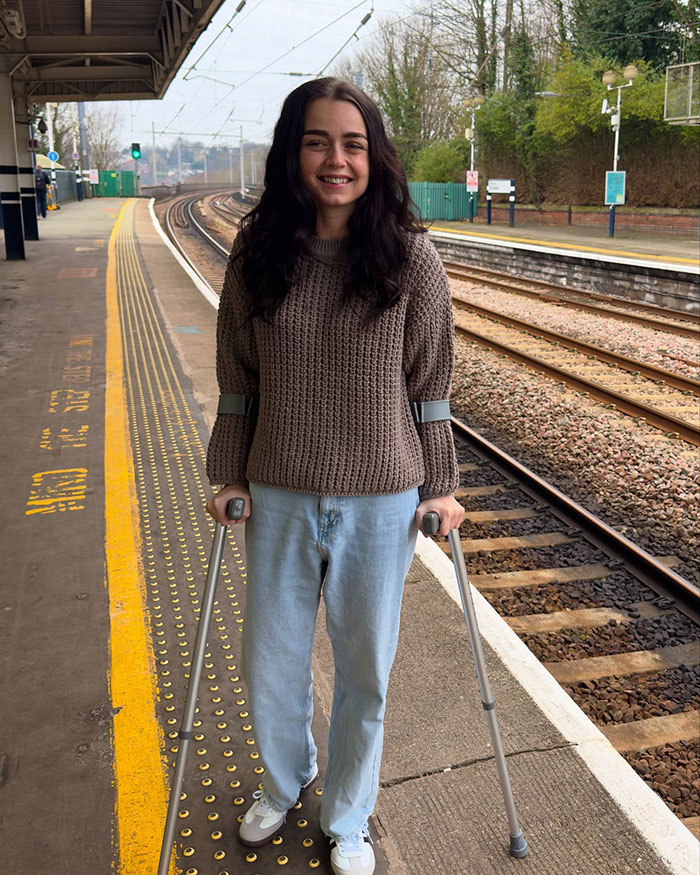 Young female pro boxer in a brown sweater using crutches, standing on a train platform near the tracks.