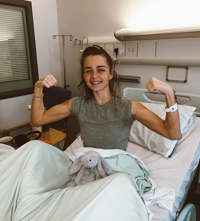 Young pro boxer Georgia O&rsquo;Connor in hospital bed flexing her arms, smiling with a stuffed bunny on the blanket.