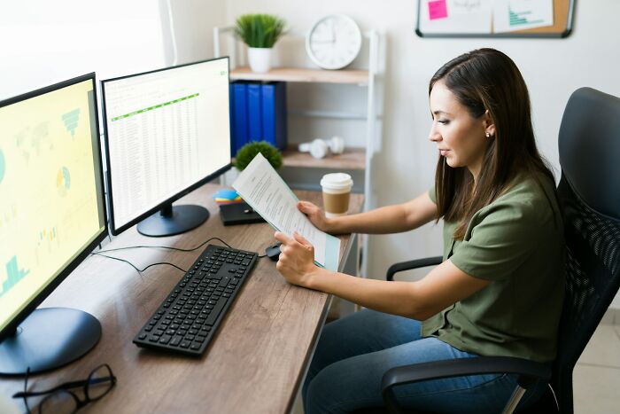 Woman analyzing documents at a desk with dual monitors, representing jobs that shouldn’t be earning as much as they do.