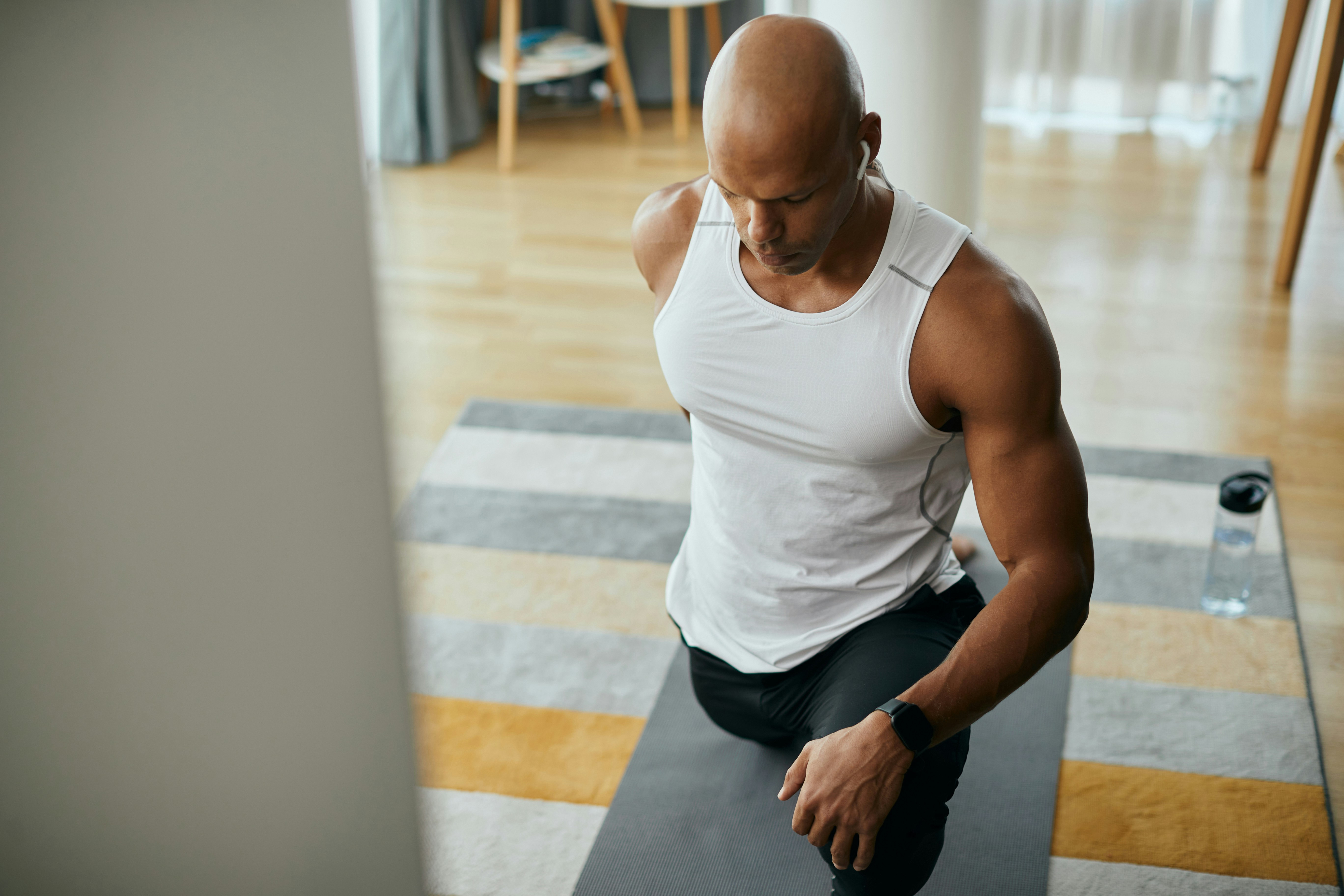 Muscular man in white tank top stretching on yoga mat at home, illustrating gym flirting and workout focus.