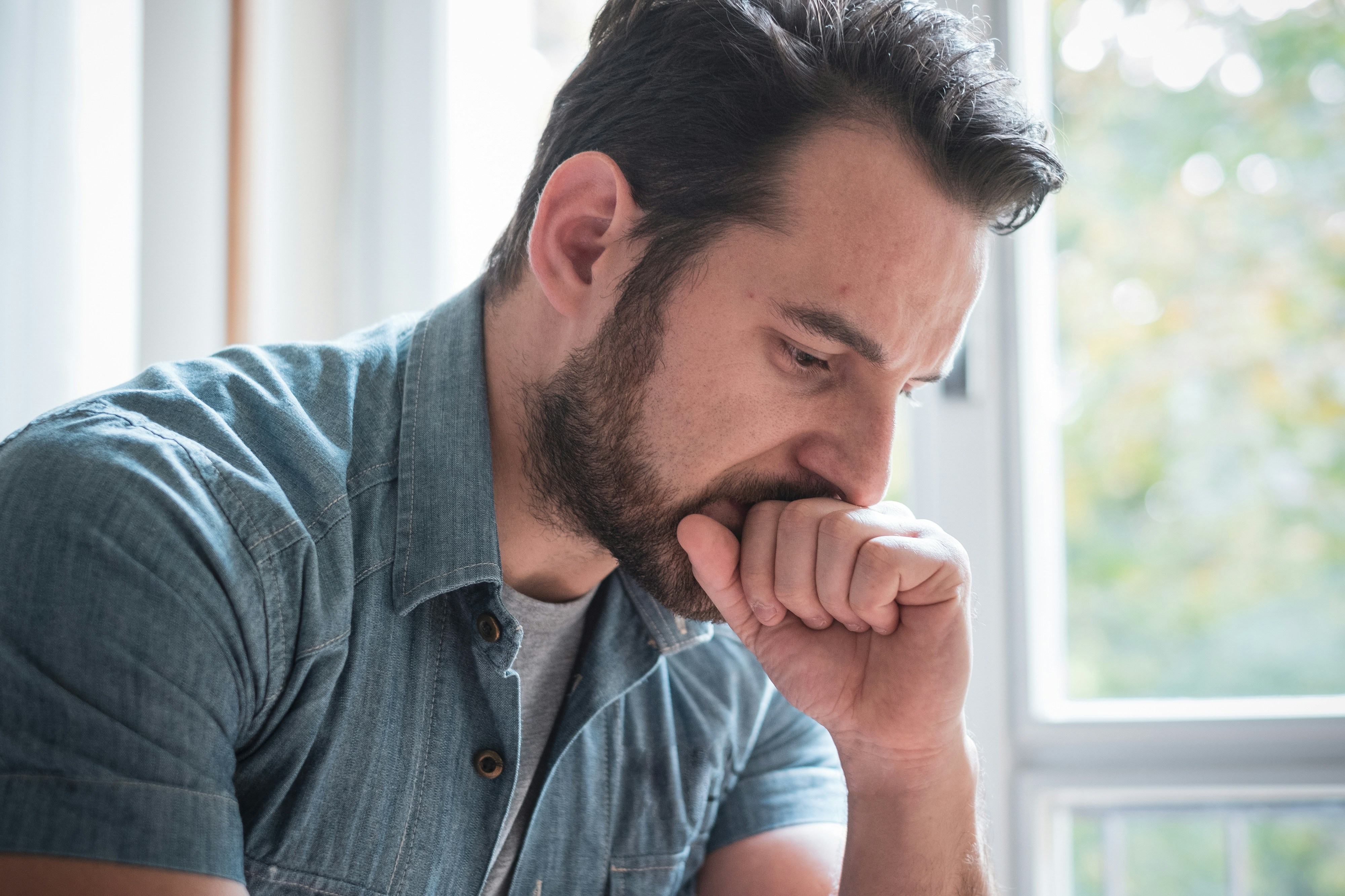 Man in denim shirt looking distressed while sitting near a window symbolizing childhood best friend favor conflict and humiliation.