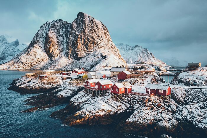 Snow-covered coastal village with red cabins and a dramatic mountain, capturing jaw-dropping natural scenery online.