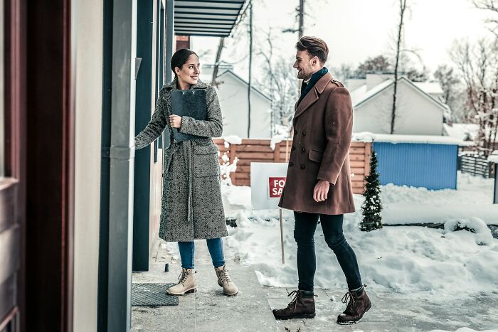 Couple dressed in winter clothes looking at a house outdoors with a for sale sign, discussing jobs earning too much.