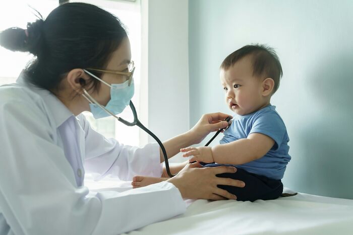 Doctor wearing mask using stethoscope to check baby sitting on exam table, illustrating hilariously bad names.