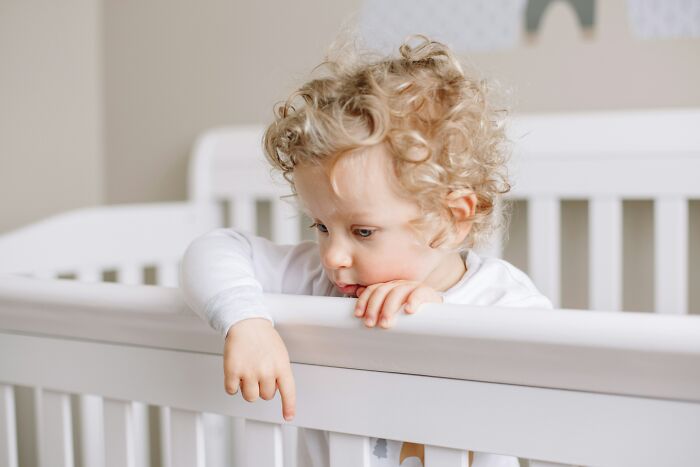 Curly-haired toddler in white pajamas leaning on crib railing, illustrating hilariously bad names in parenting choices.